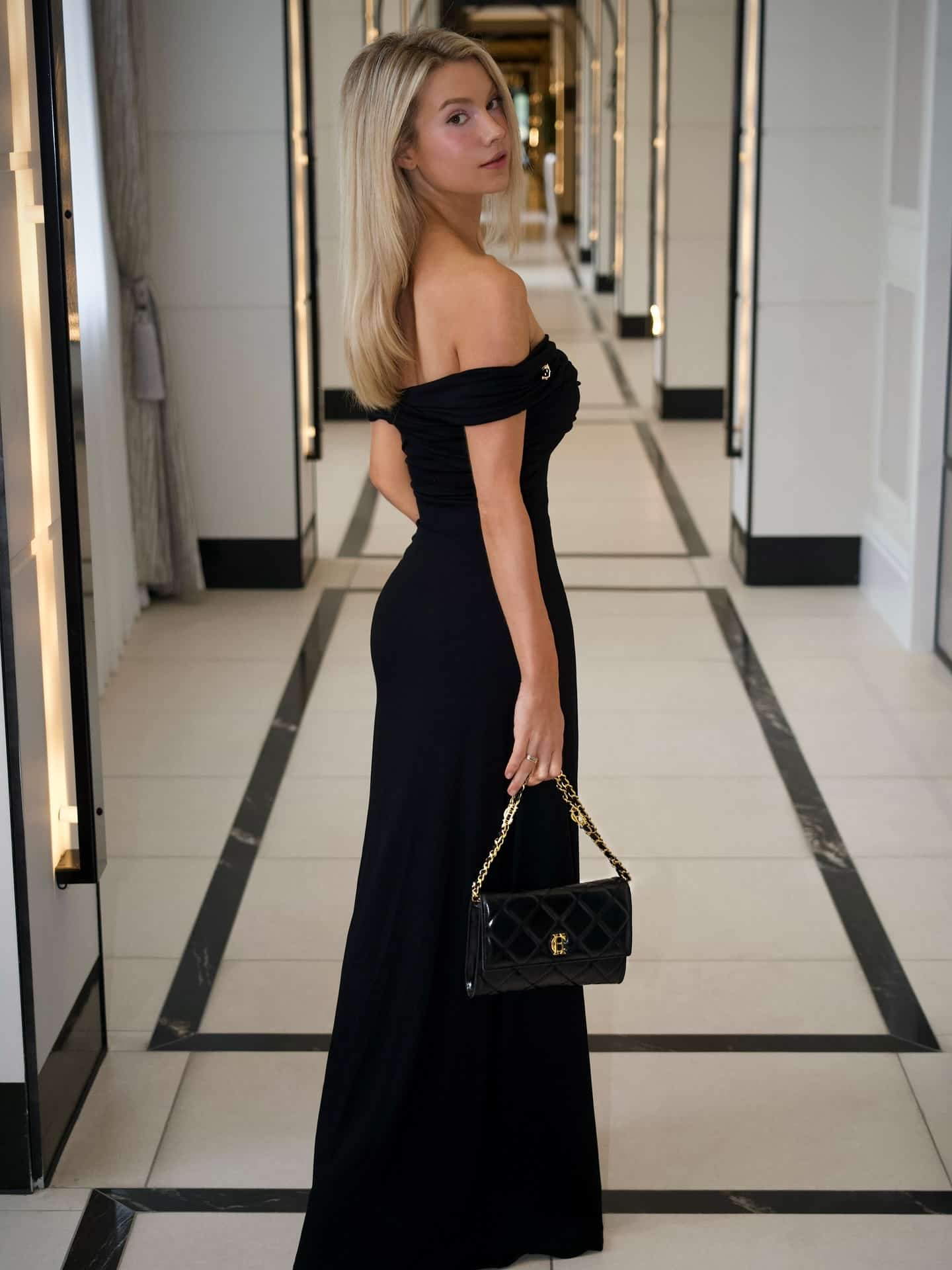 Lucy Robson defines high-end luxury fashion in a timeless black off-the-shoulder evening gown elegantly accessorized with a quilted designer handbag.